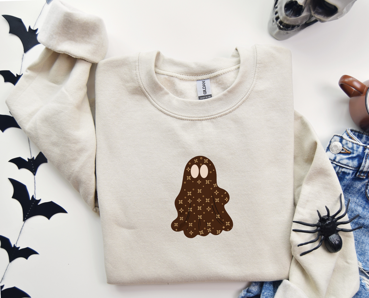 Folded beige sweatshirt with a brown ghost design on a white surface with Halloween decorations.