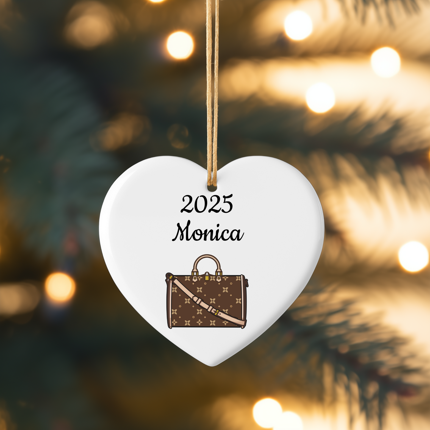 Heart-shaped ornament with '2025 Monica' text and a suitcase design, hanging against a blurred Christmas tree background.