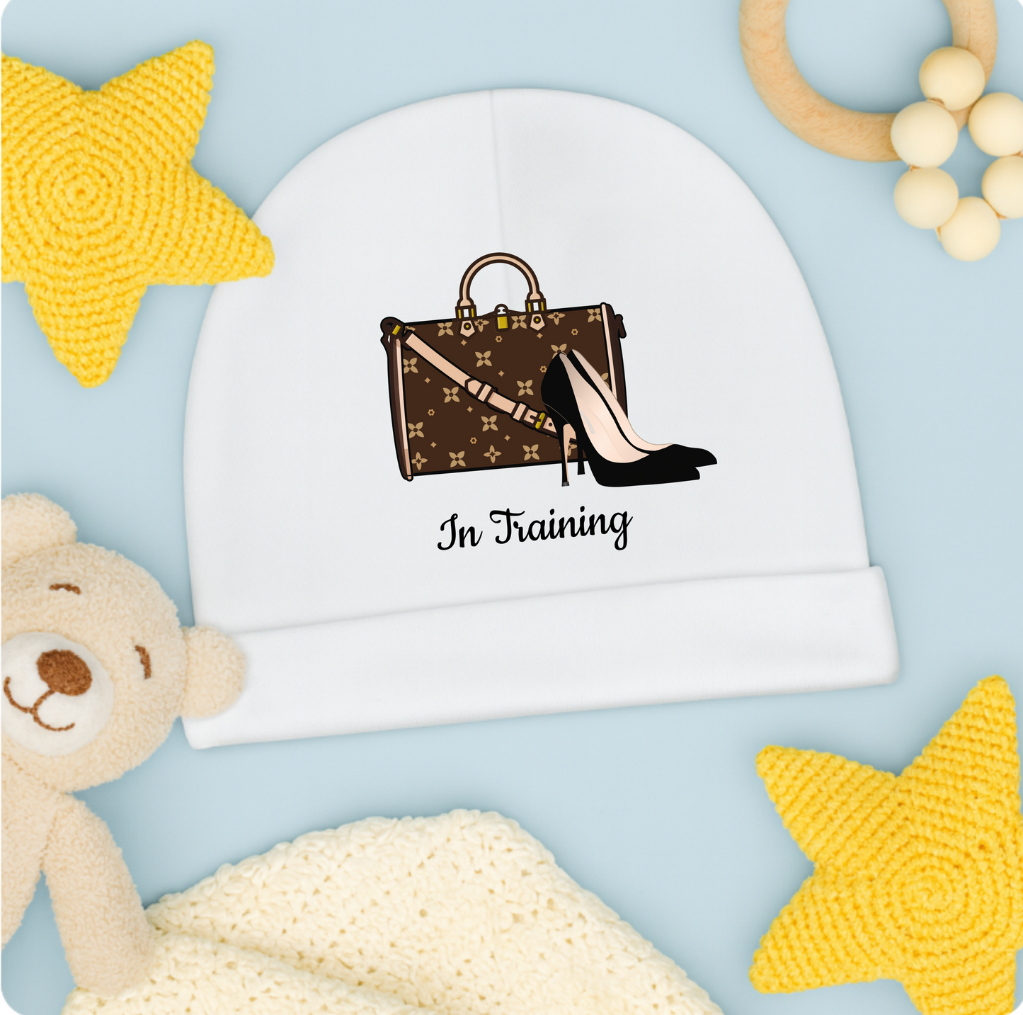 White beanie with a handbag and shoe design and 'In Training' text, surrounded by toys on a light blue background.