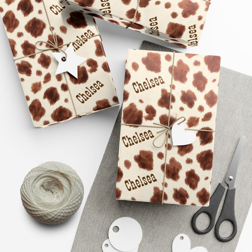 Gift boxes with a cow print pattern and 'Chelsea' branding, accompanied by yarn and scissors on a white surface.