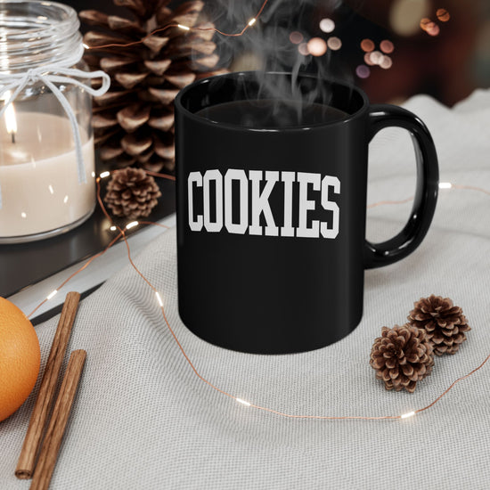 Black mug with 'COOKIES' text on a table with decorative items including a candle, oranges, and pine cones.