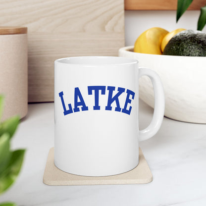 White mug with 'LATKE' printed in blue on a white surface with a plant and container in the background.