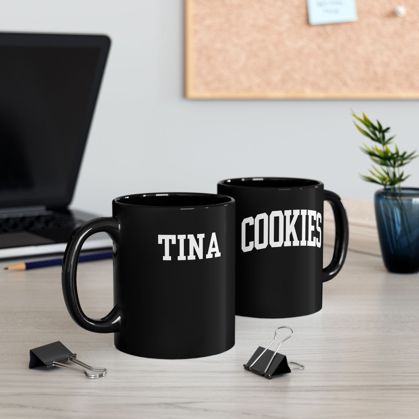Two black mugs labeled 'TINA' and 'COOKIES' on a desk with office supplies.