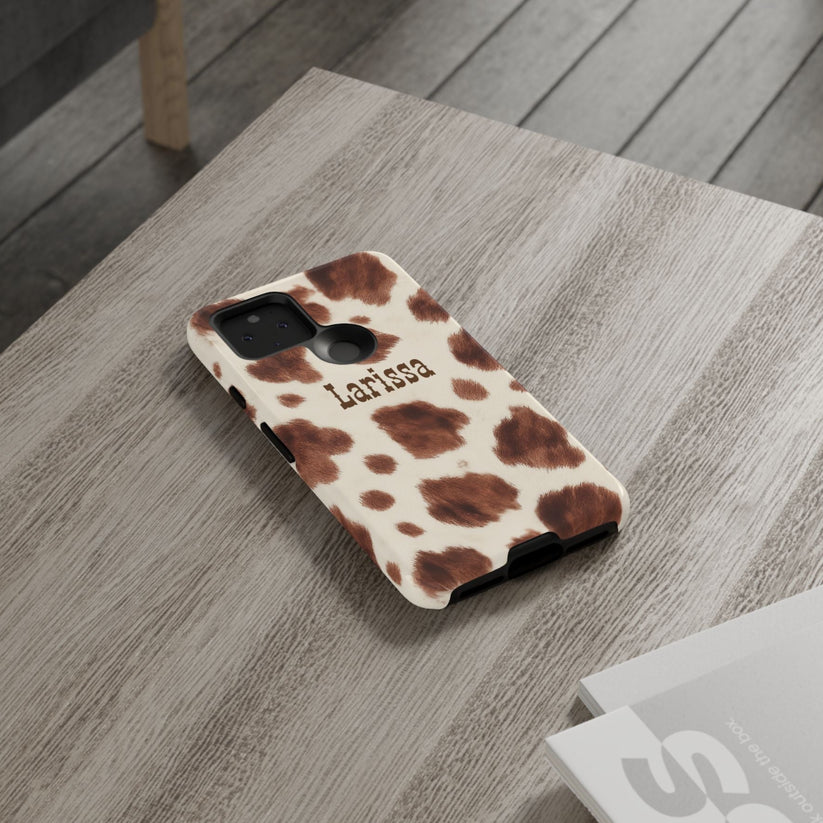 Cow print phone case with 'Larissa' branding on a wooden surface