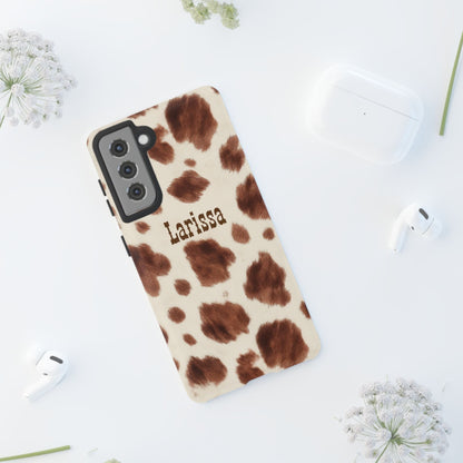Cow print phone case with 'Larissa' branding on a white surface with flowers.