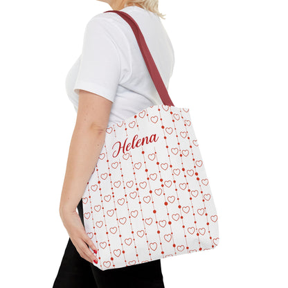 Person holding a white tote bag with red heart patterns and 'Helena' text.