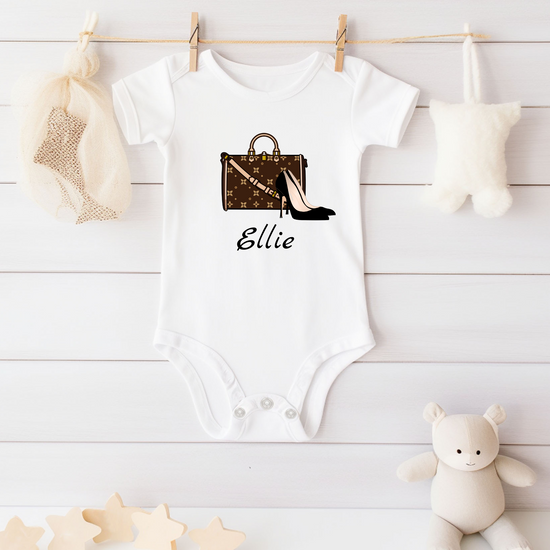 White baby onesie with fashion-themed design and name 'Ellie' on a wooden surface.