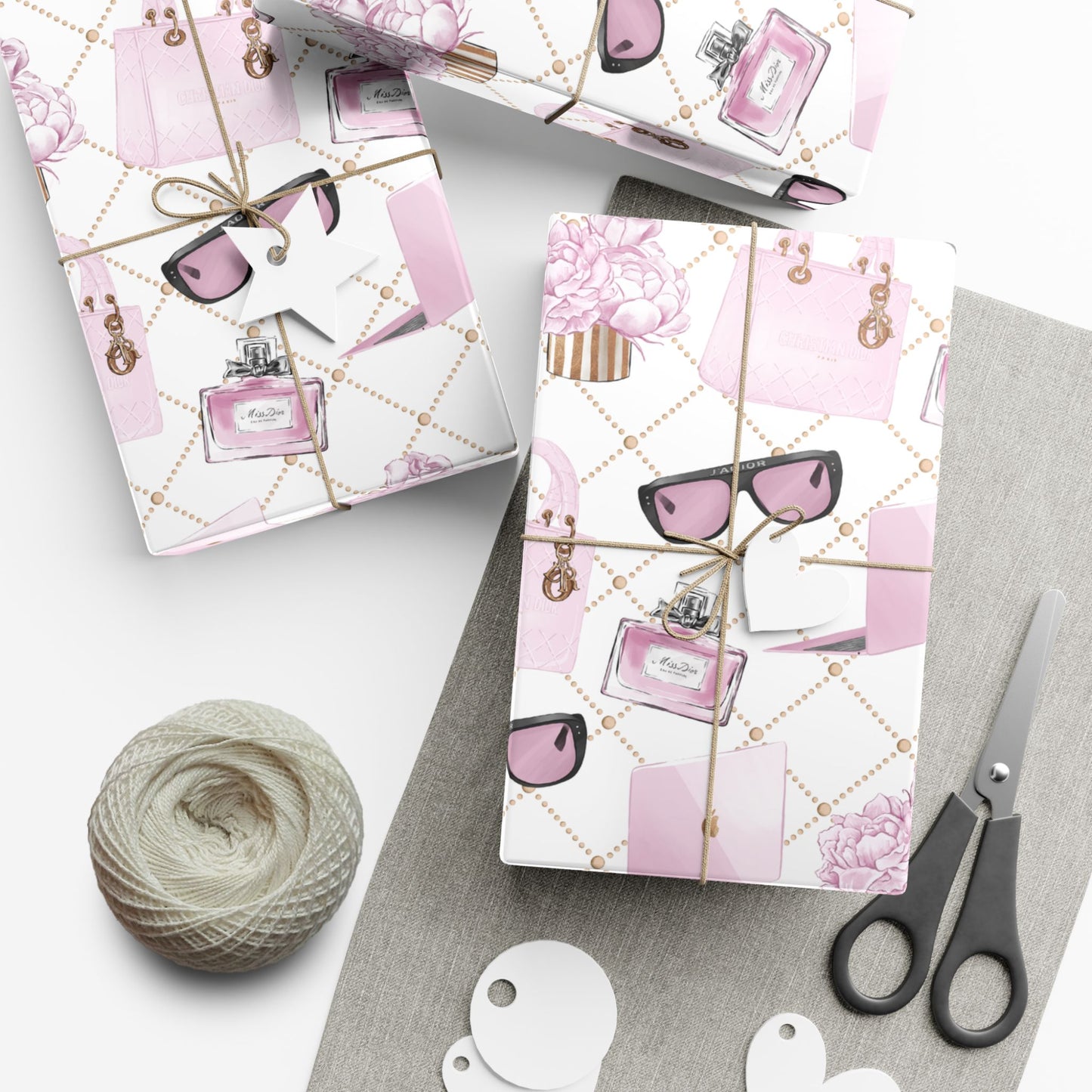 Gift wrap paper with pink and white fashion accessory design featuring sunglasses and perfume bottles, tied with string, on a white surface with scissors and yarn.