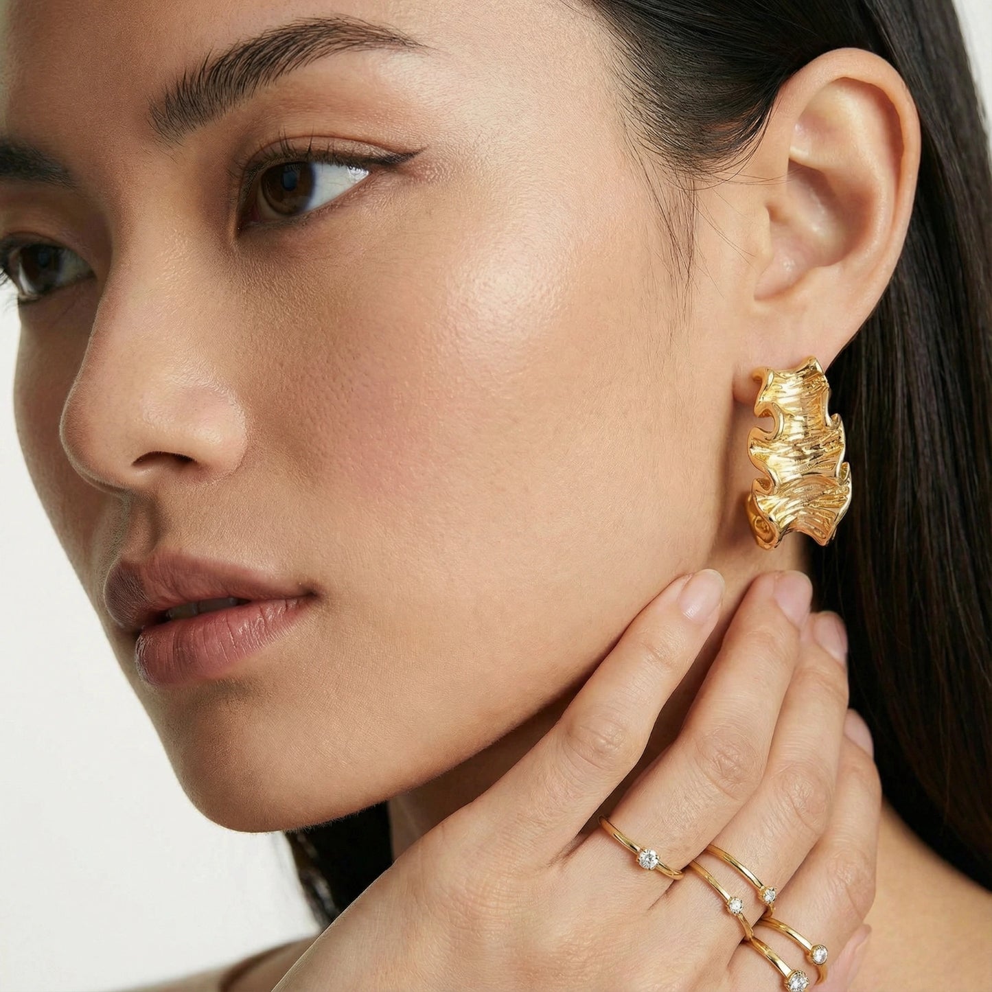 Close-up of a woman wearing gold  ruffle earrings and rings on a white background