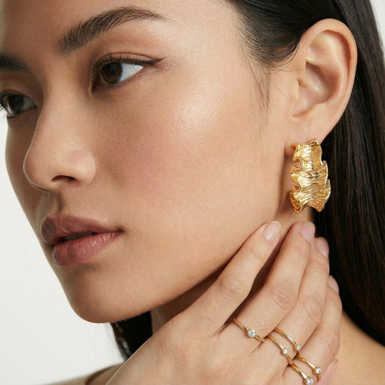 Close-up of a woman wearing gold  ruffle earrings and rings on a white background