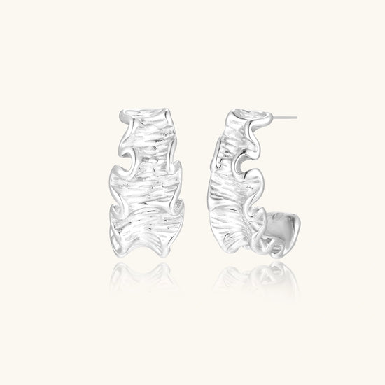 White gold plated sculptural hoop earrings with organic wavy edges and reflective metallic finish on cream background