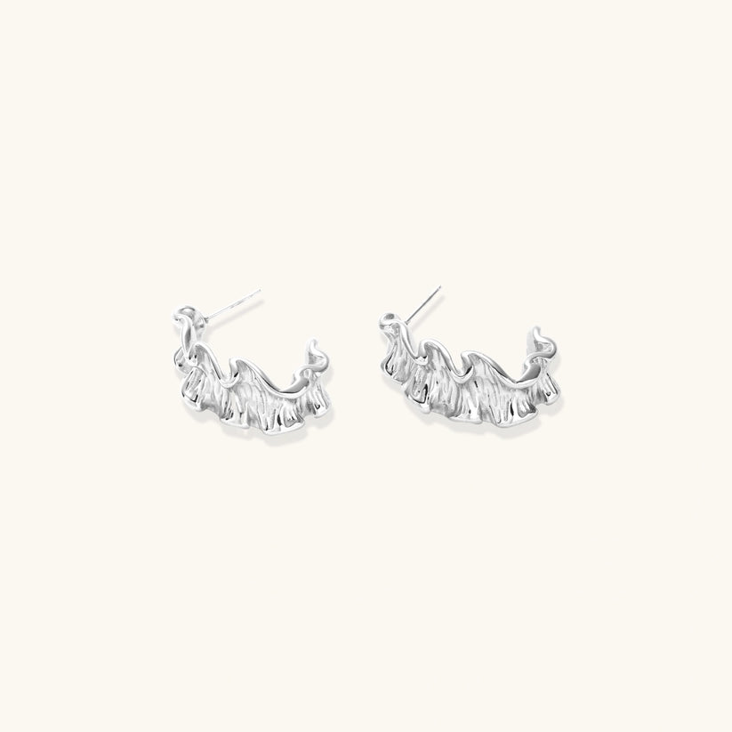 White gold plated sculptural hoop earrings with organic wavy edges and reflective metallic finish on cream background