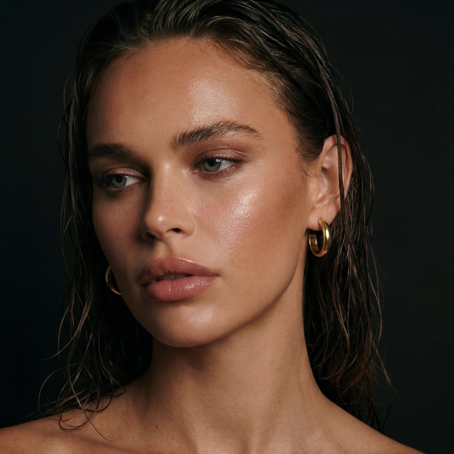 Close-up of a woman with wet hair wearing gold hoop earrings against a dark background