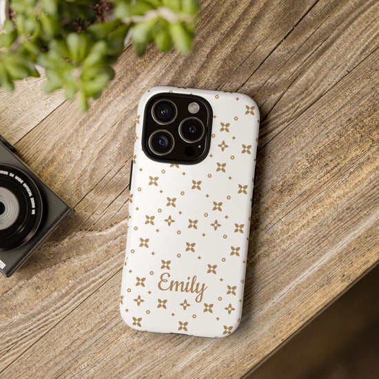Phone case with gold pattern and 'Emily' engraving on a wooden surface with a camera and plant.