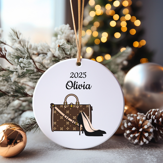 Decorative ornament with '2025 Olivia' text, handbag, and high heel illustration, surrounded by Christmas decorations.