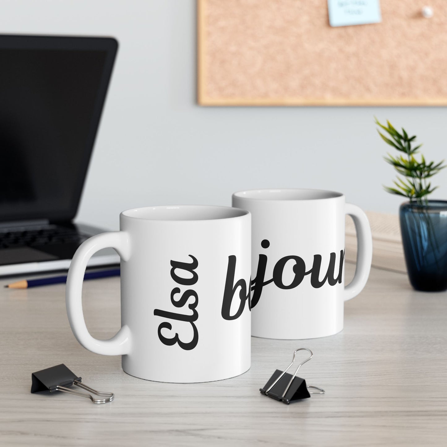 Two white mugs with 'Elsa' and 'bijou' text on a desk with office supplies.