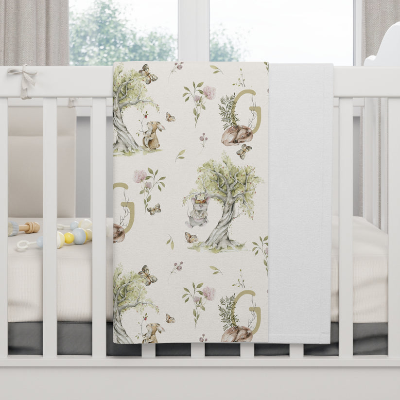 Crib with a Soft plush baby blanket, cream background with watercolor illustrations of a bunny and mouse.  Shown on a white, crib in a bright room. Personalized with a monogram letter.