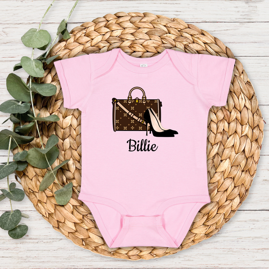 Pink baby onesie with a design and name 'Billie' on a woven mat with eucalyptus leaves.