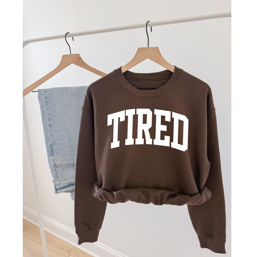 Brown sweatshirt with 'TIRED' text on a hanger against a white wall.