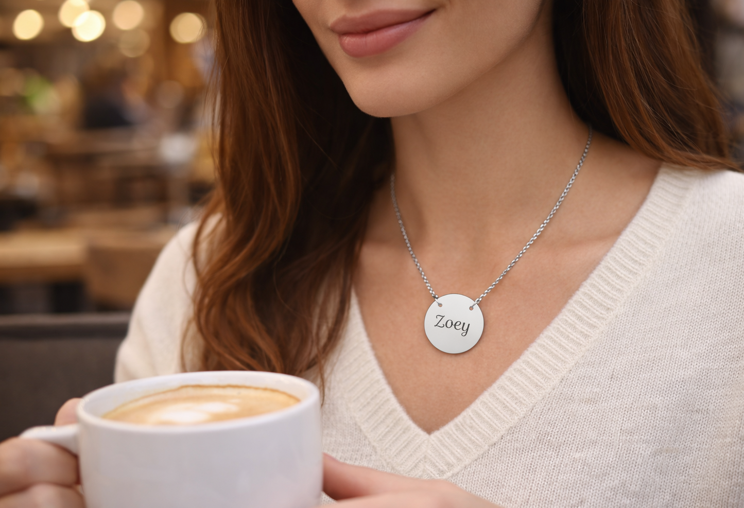 Woman holding a cup of coffee wearing a necklace with a 'Zoey' pendant in a blurred cafe setting