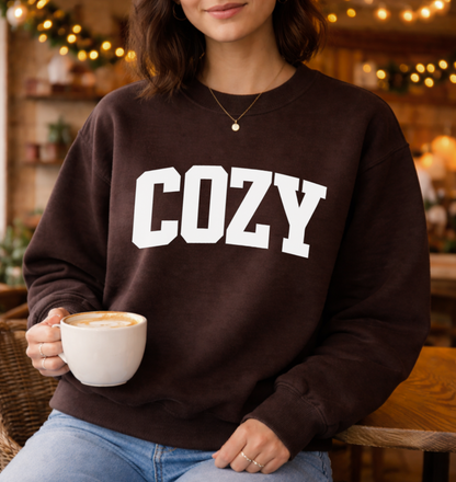 Person wearing a brown sweatshirt with 'COZY' text, holding a cup of coffee in a cozy setting.
