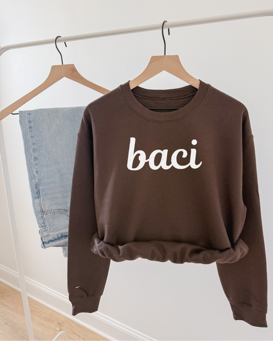 Brown sweatshirt with 'baci' text on a hanger against a white wall.
