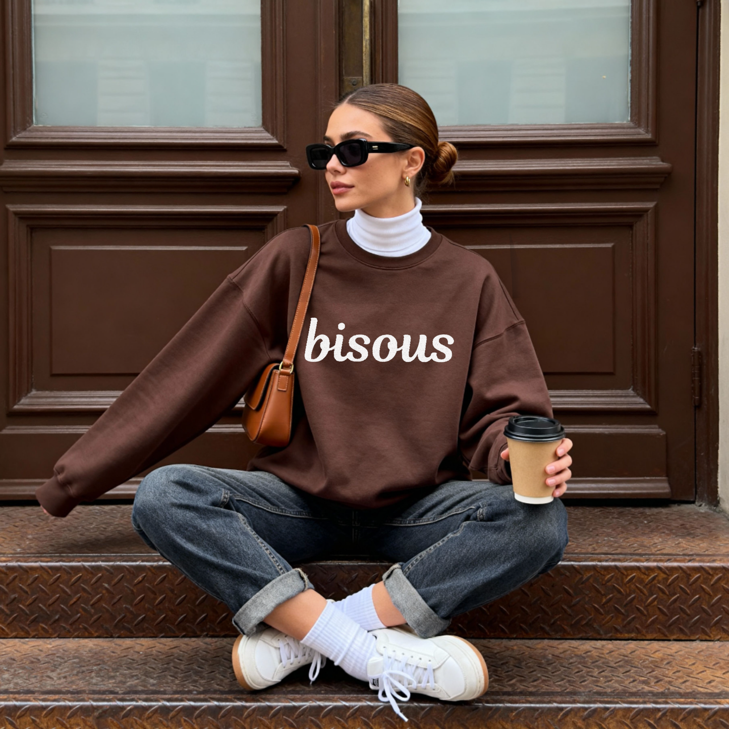 Woman sitting on steps wearing a brown sweatshirt with white embroidery text 'bisous' , holding a coffee cup.