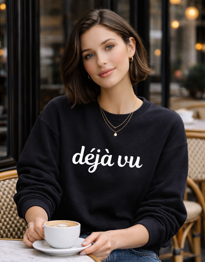 Woman wearing a black sweatshirt with 'déjà vu' embroidered text, holding a coffee cup in an outdoor cafe setting.
