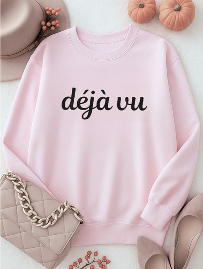 Pink sweatshirt with 'déjà vu' embroidered text on a white surface with accessories.
