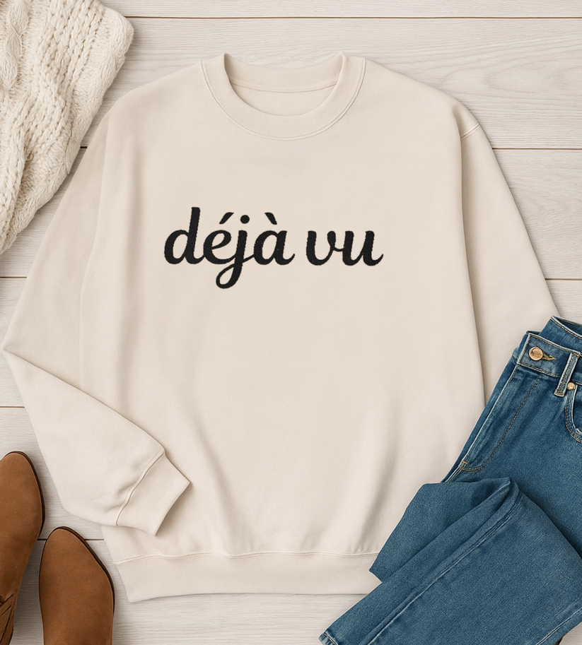Beige sweatshirt with 'déjà vu' text on a wooden surface with jeans and shoes.