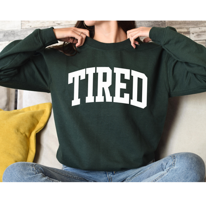 Person wearing a dark green sweatshirt with 'TIRED' printed on it, sitting on a couch.