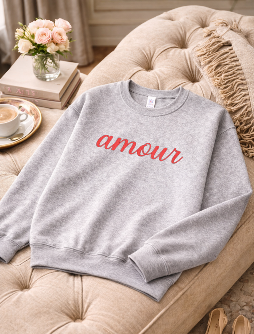 Gray sweatshirt with 'amour' text on a beige couch with a cup of coffee and flowers in the background.