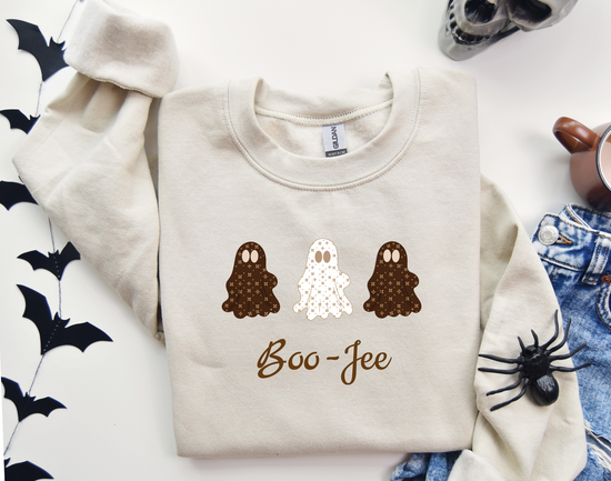 Beige sweatshirt with Halloween-themed design featuring ghosts and 'Boo-Jee' text, placed on a white surface with decorative items.
