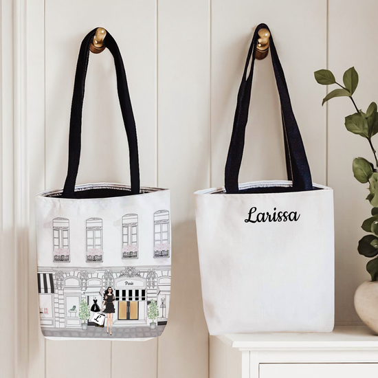 Two tote bags with black handles on a white surface, one featuring an illustration and the other with 'Larissa' text.
