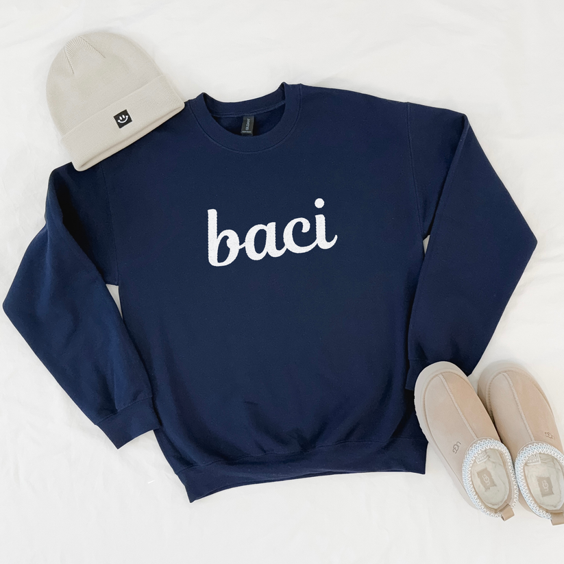 Navy blue sweatshirt with 'baci' text, beige beanie, and shoes on a white background