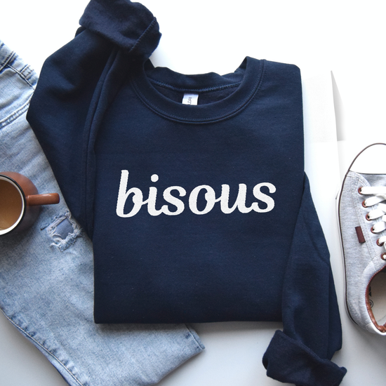 Navy blue sweatshirt with 'bisous' embroidered  text in white on a white surface with denim jeans and a shoe.