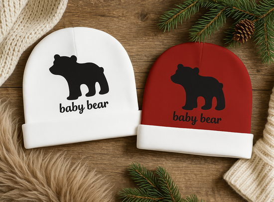 Two baby hats with bear designs and 'baby bear' text on a wooden surface with Christmas decorations.
