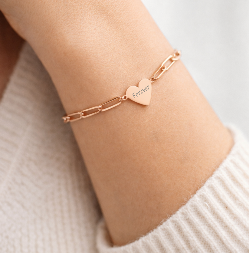 Rose gold bracelet with a heart-shaped tag on a wrist against a neutral background