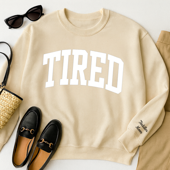 Beige sweatshirt with 'TIRED' text, black loafers, sunglasses, and a handbag on a white surface