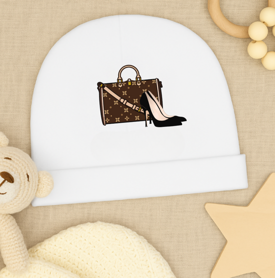 White baby beanie with a brown handbag and black high heel design, placed on a beige surface with a teddy bear and rattle.
