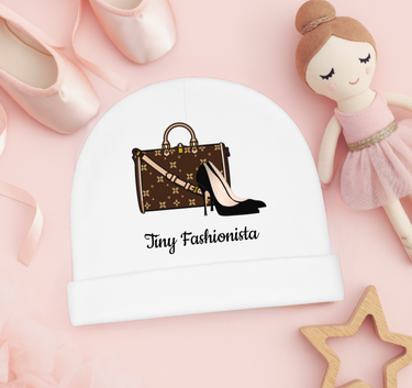 White baby hat with 'Tiny Fashionista' design featuring a handbag and high heels, on a pink background with a doll and shoes.