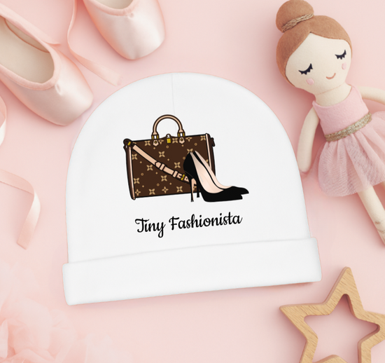 White baby hat with 'Tiny Fashionista' design featuring a handbag and high heels, on a pink background with a doll and shoes.