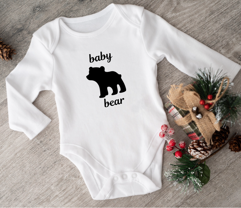 White baby onesie with 'baby bear' print on a wooden surface with decorative items.