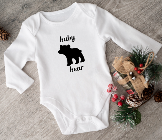 White baby onesie with 'baby bear' print on a wooden surface with decorative items.