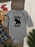 Gray baby onesie with 'baby bear' print on a wooden surface with decorative items.