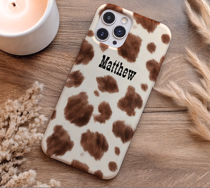 Phone case with cow print pattern and 'Matthew' text on a wooden surface.