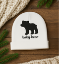 White baby hat with black bear design and 'baby bear' text on a wooden surface with greenery and beige fabric.
