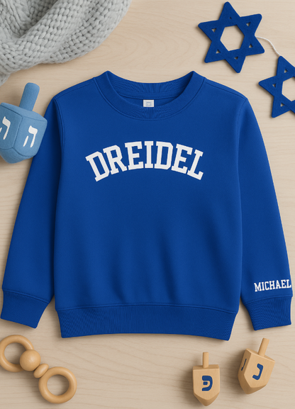 Blue sweatshirt with 'DREIDEL' printed on it, surrounded by wooden dreidels and a Star of David, on a wooden surface.