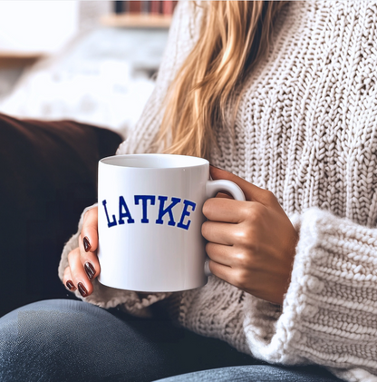 Person holding a white mug with 'LATKE' printed on it, wearing a beige sweater.