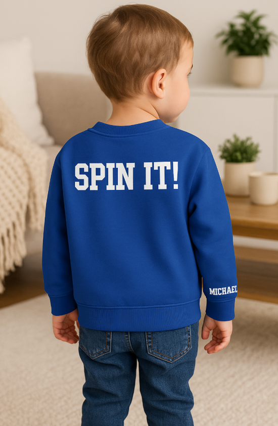 Child wearing a blue sweatshirt with 'SPIN IT!' text in a home setting